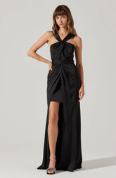 A woman is wearing a sleeveless black evening gown with a knotted design at the bust and waist. The dress features wide shoulder straps that cross over at the shoulders and a high slit on the left side, revealing her leg. She is also wearing black high-heeled sandals with embellishments.